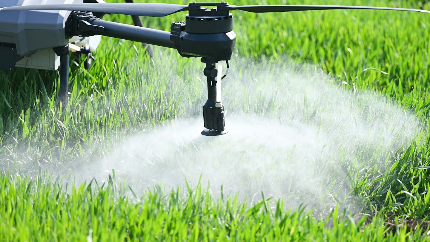 Un drone agricolo grigio spruzza nebbia su un campo di colture verdi.